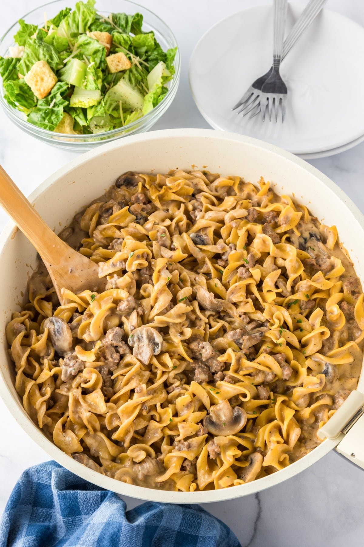 A pan full of ground beef stroganoff being scooped with a spoon, with salad and plates nearby.