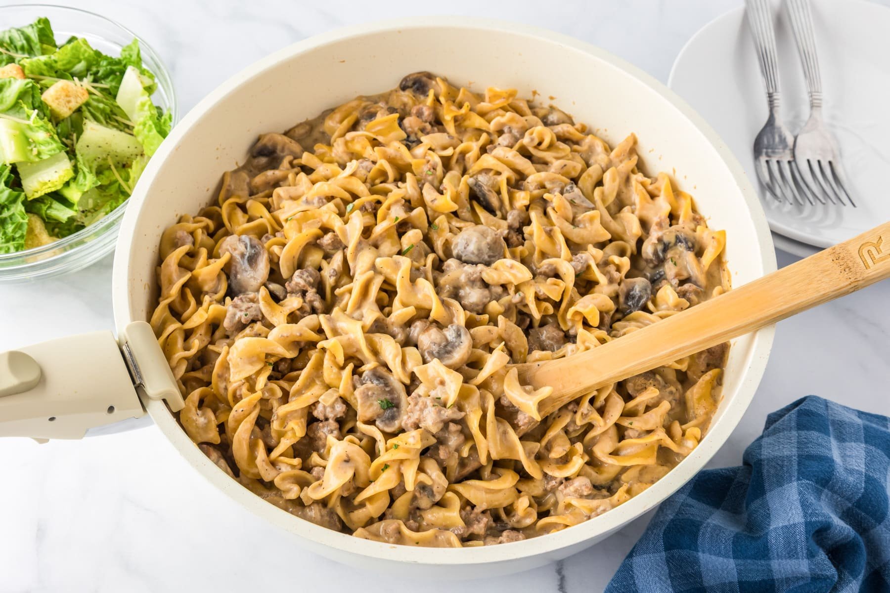A pan filled with creamy beef stroganoff made with egg noodles and mushrooms being stirred with a wooden spoon on a counter.