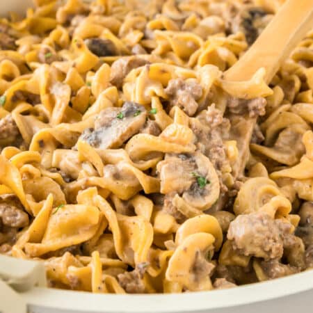 Close-up of creamy ground beef stroganoff with egg noodles, ground beef, mushrooms, and sauce being stirred with a wooden spoon.