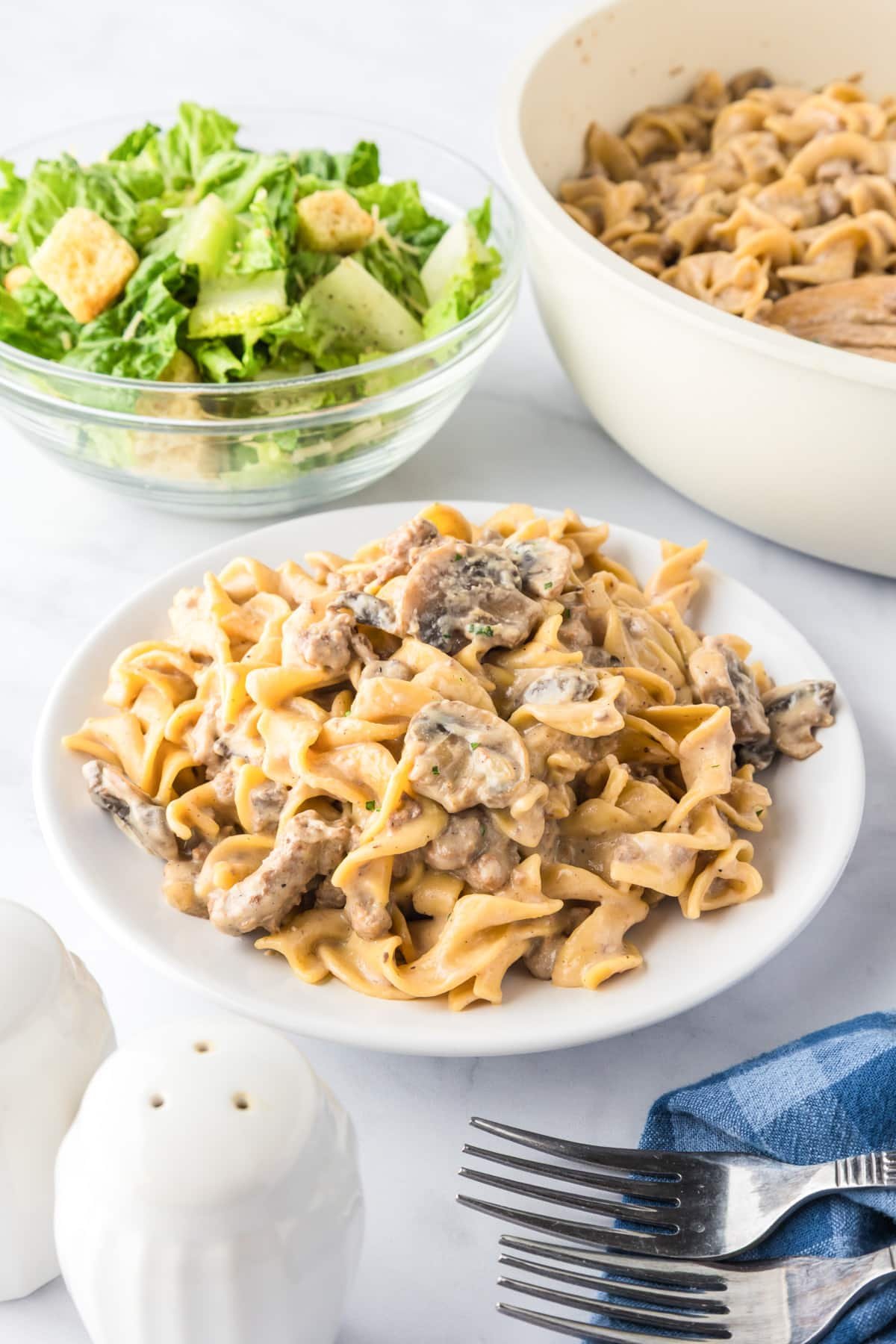 A plate of creamy ground beef stroganoff with the rest of the pan full and a small salad nearby.