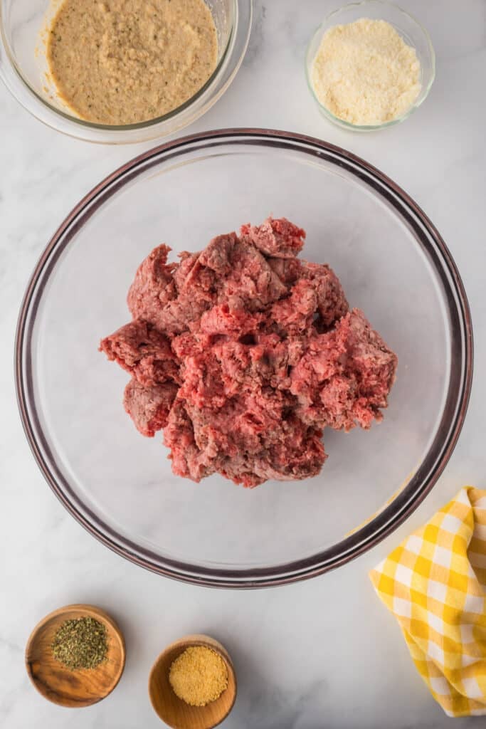 A large glass bowl of raw ground beef is surrounded by bowls containing breadcrumbs combined with milk, grated parmesan cheese, and spices for Italian meatloaf.