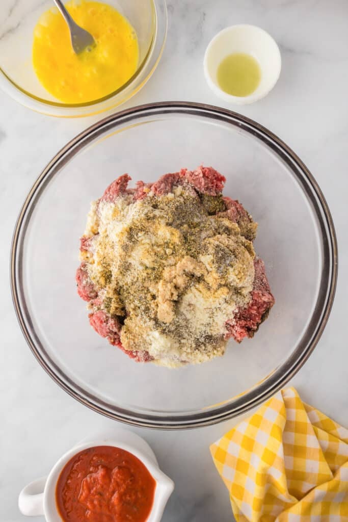 A glass bowl with ground beef, bread crumbs, grated cheese, and seasonings, surrounded by a bowl of beaten eggs, a small bowl of oil and a cup of marinara.