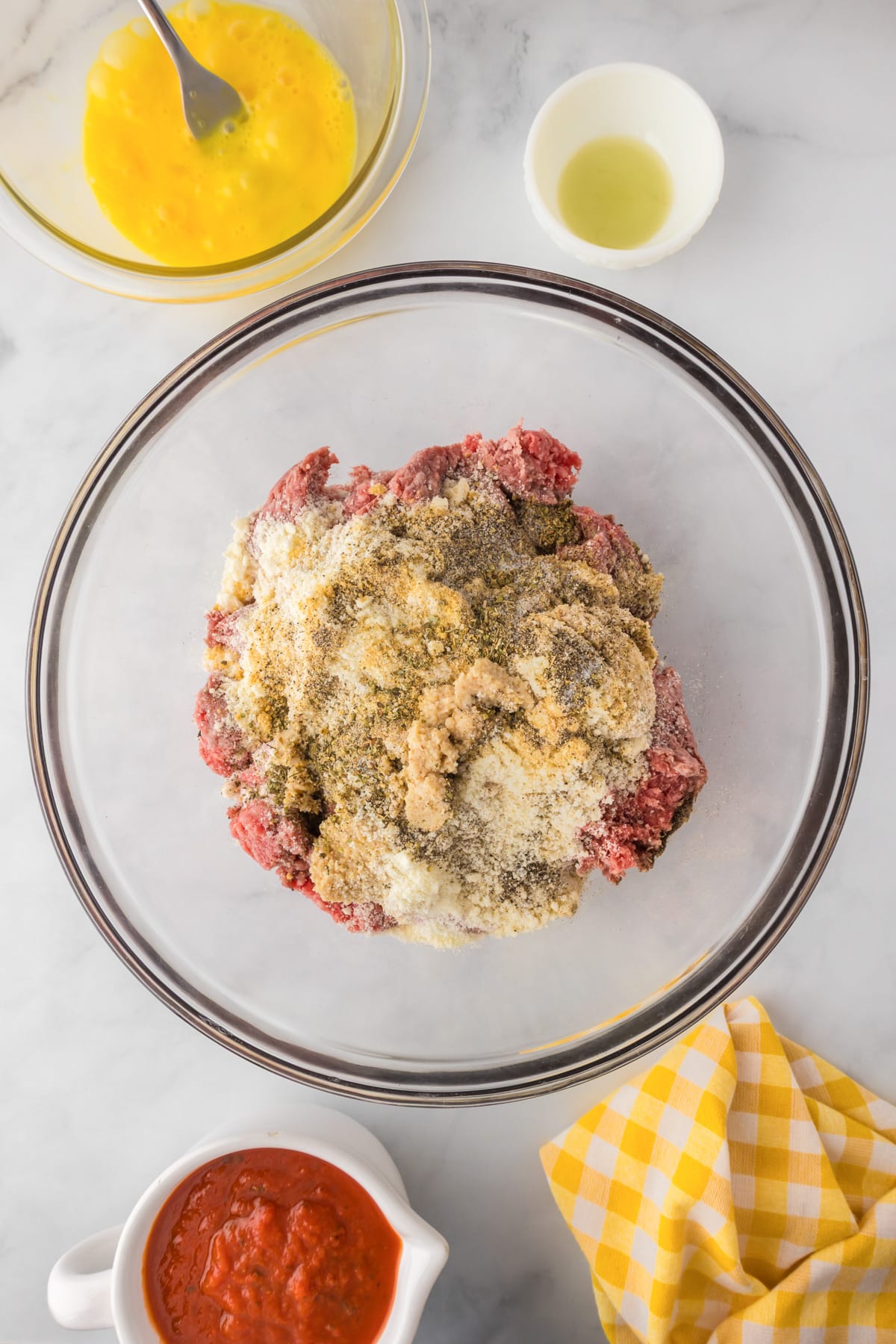 A glass bowl with ground beef, bread crumbs, grated cheese, and seasonings, surrounded by a bowl of beaten eggs, a small bowl of oil and a cup of marinara.