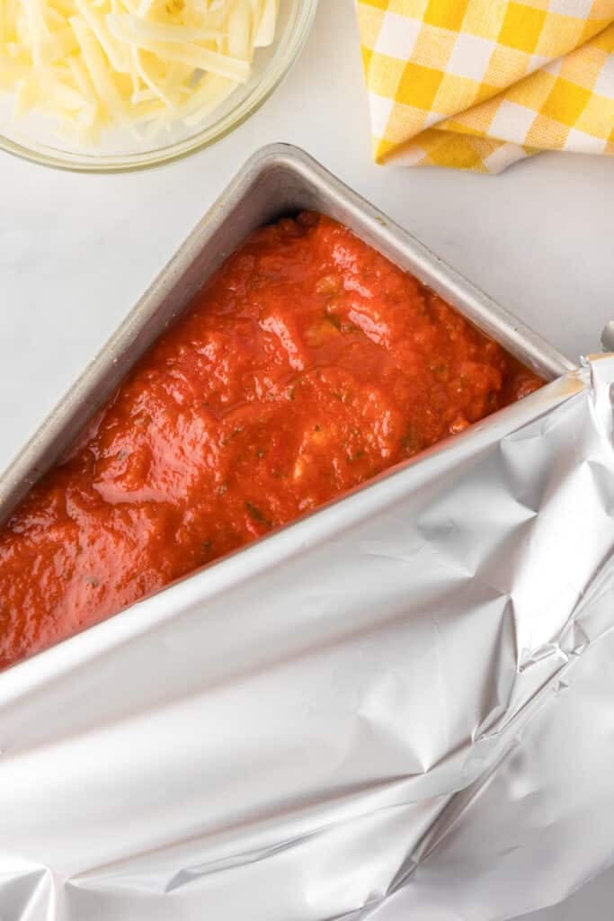 A rectangular baking pan with tomato sauce and full of Italian meatloaf being covered with aluminum foil.