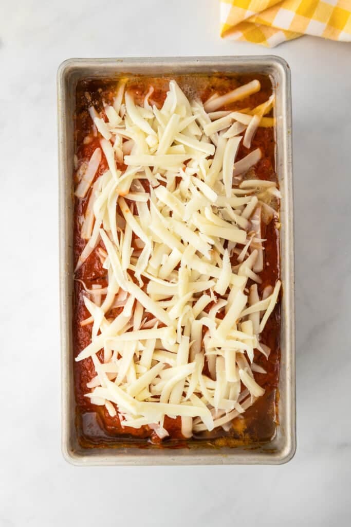 A rectangular metal pan filled with baked Italian meatloaf being topped with mozzarella cheese.