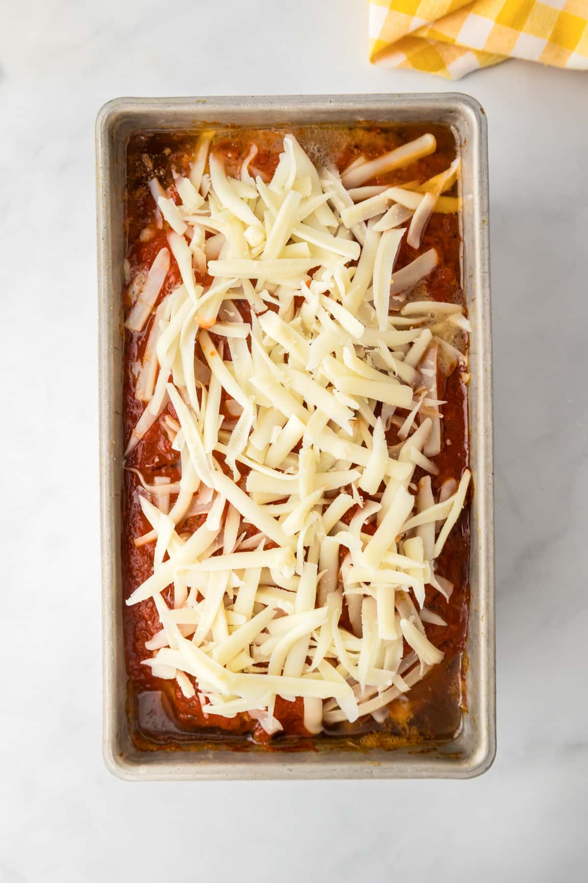 A rectangular metal pan filled with baked Italian meatloaf being topped with mozzarella cheese.