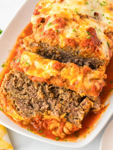 Square view of a sliced Italian meatloaf topped with melted cheese and tomato sauce is served on a white platter with sauce around it.