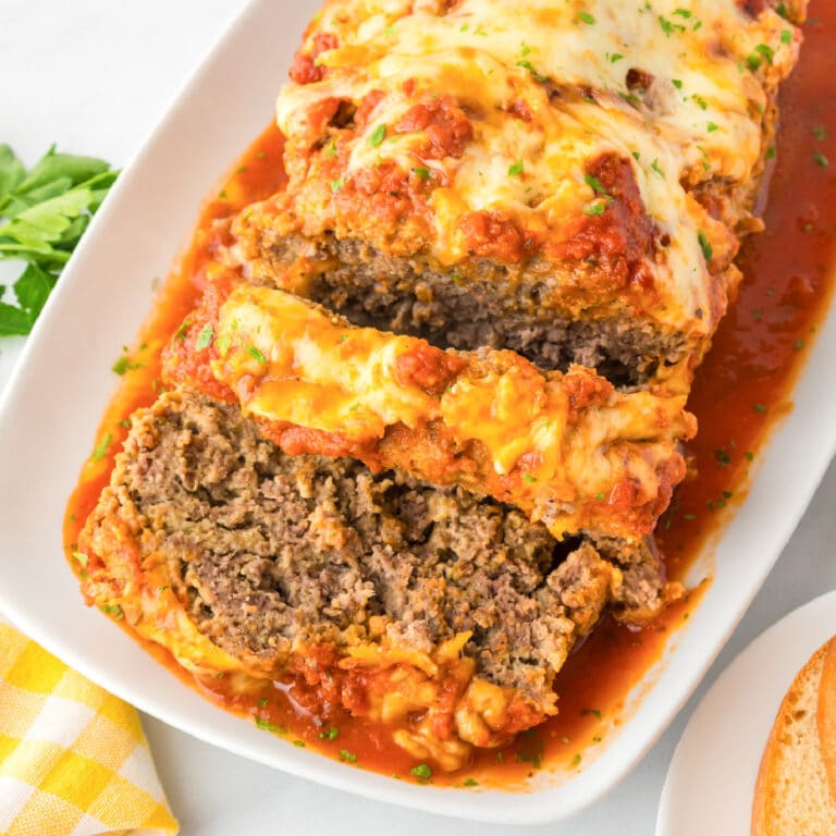 Square view of a sliced Italian meatloaf topped with melted cheese and tomato sauce is served on a white platter with sauce around it.