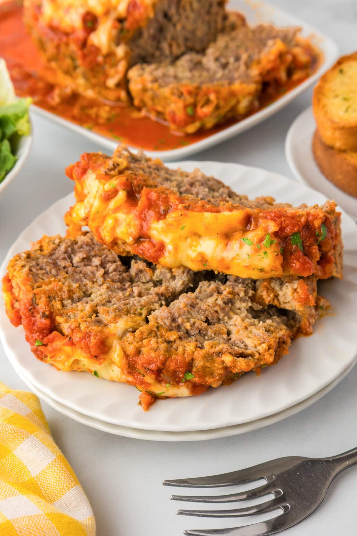 Two slices of Italian meatloaf topped with tomato sauce and melted cheese on a plate with more on a platter in the background.