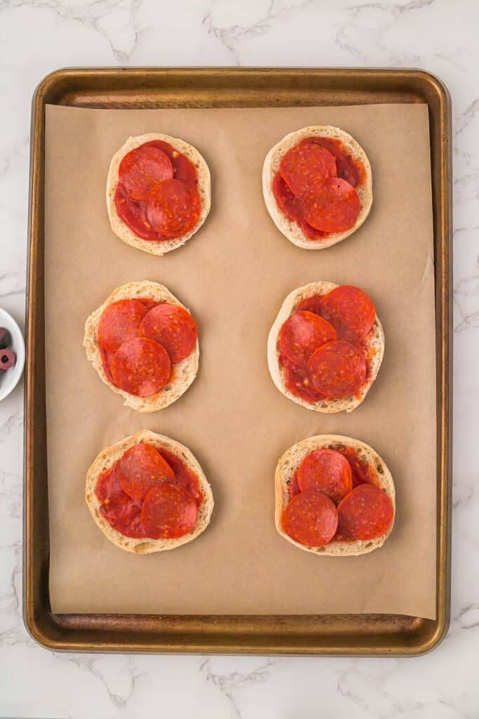 A baking sheet lined with parchment paper holds six English muffin halves transformed into mini mummy pizzas, each topped with pizza sauce and pepperoni slices.