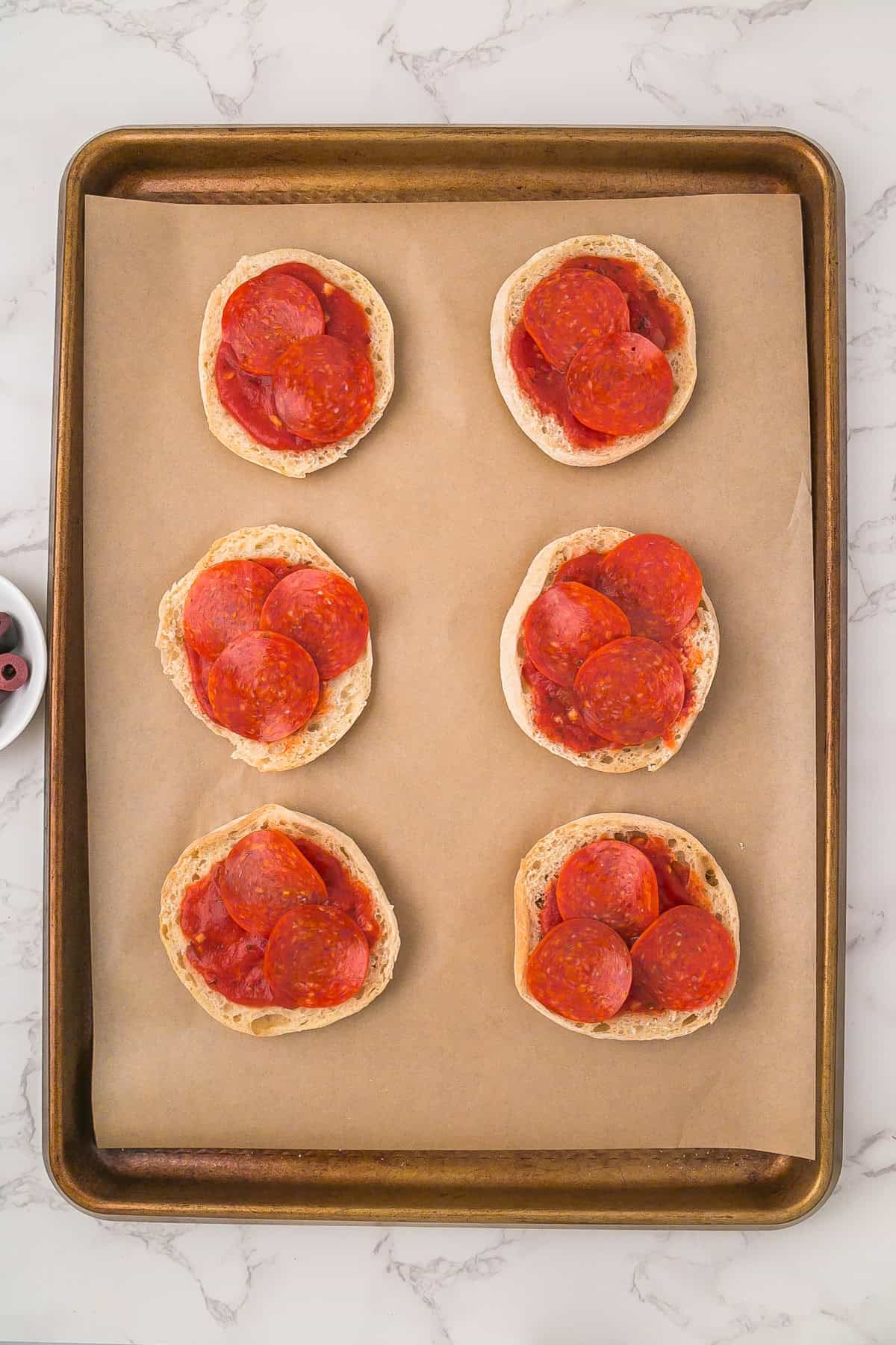 A baking sheet lined with parchment paper holds six English muffin halves transformed into mini mummy pizzas, each topped with pizza sauce and pepperoni slices.