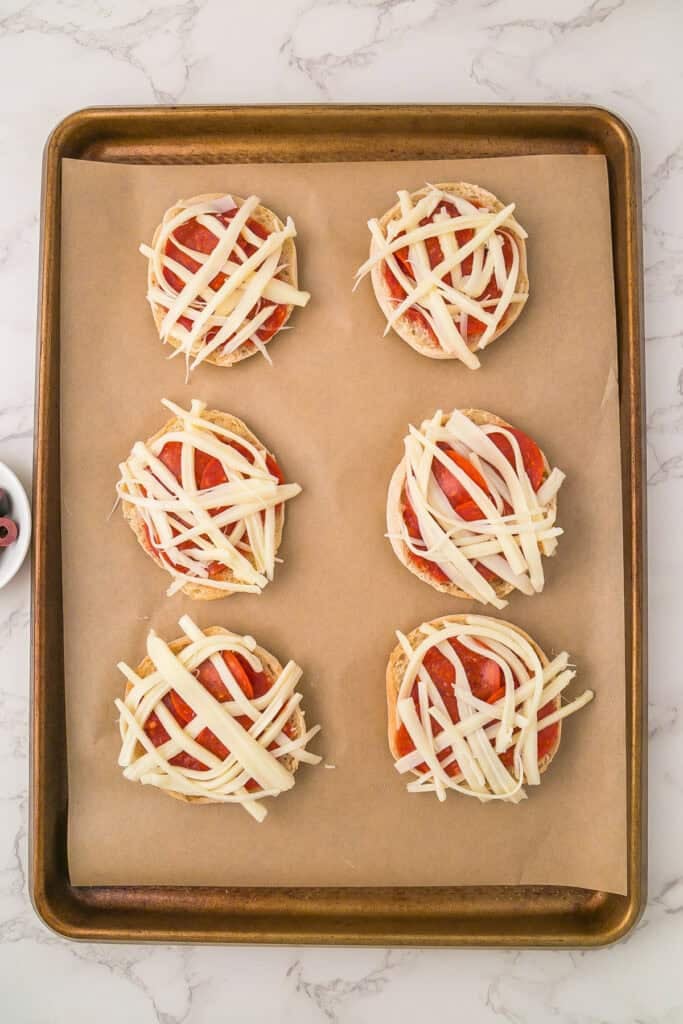 A baking sheet with six mini mummy pizzas—English muffin halves topped with tomato sauce, pepperoni slices, and strips of mozzarella cheese—rests on parchment paper.