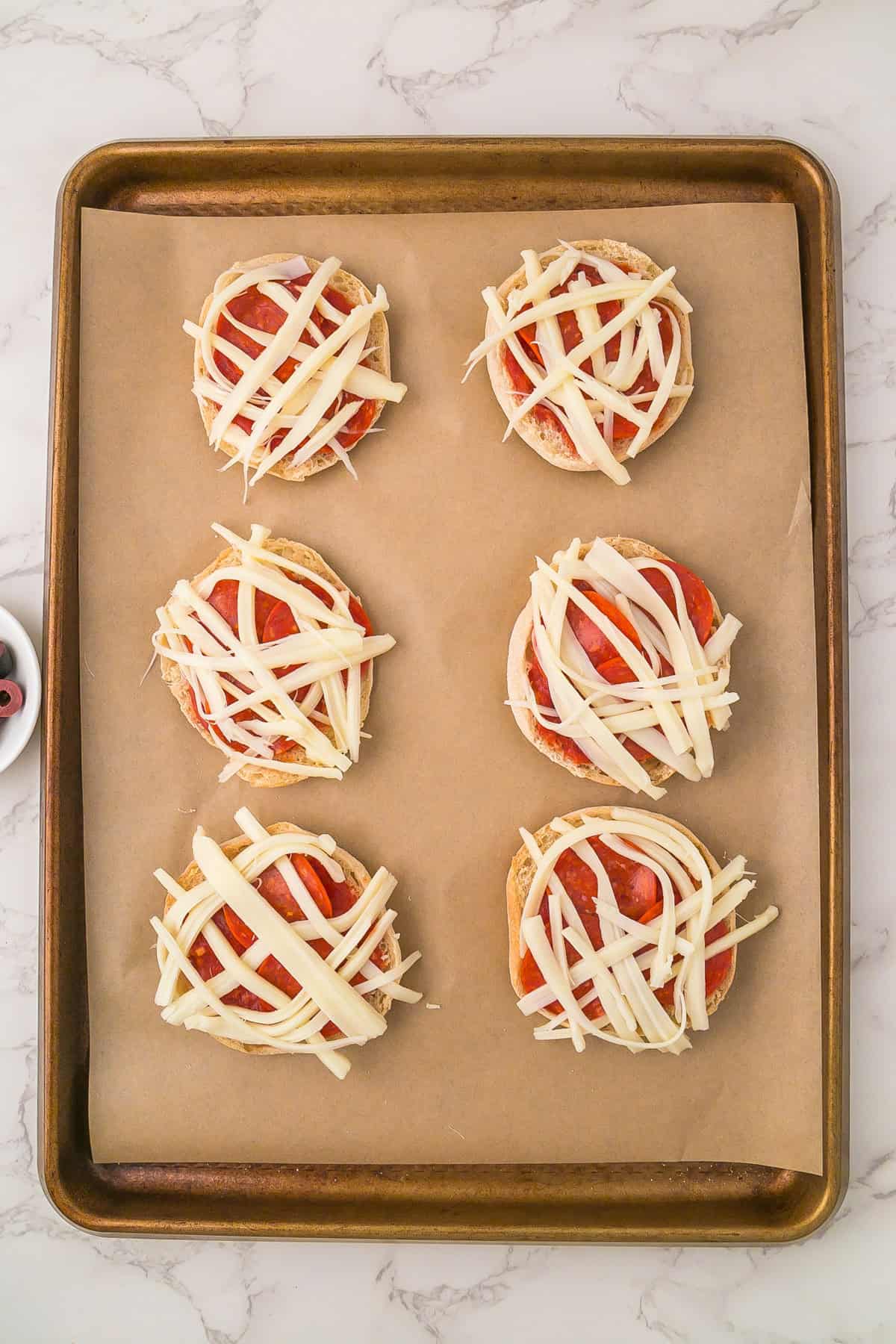 A baking sheet with six mini mummy pizzas—English muffin halves topped with tomato sauce, pepperoni slices, and strips of mozzarella cheese—rests on parchment paper.