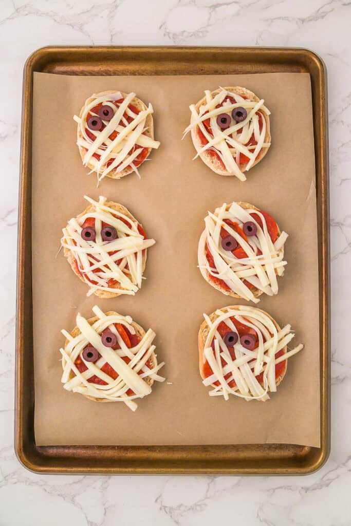 Six unbaked mini mummy pizzas made from English muffins are decorated with strips of cheese and olive slice eyes, all arranged on a baking sheet lined with parchment paper.
