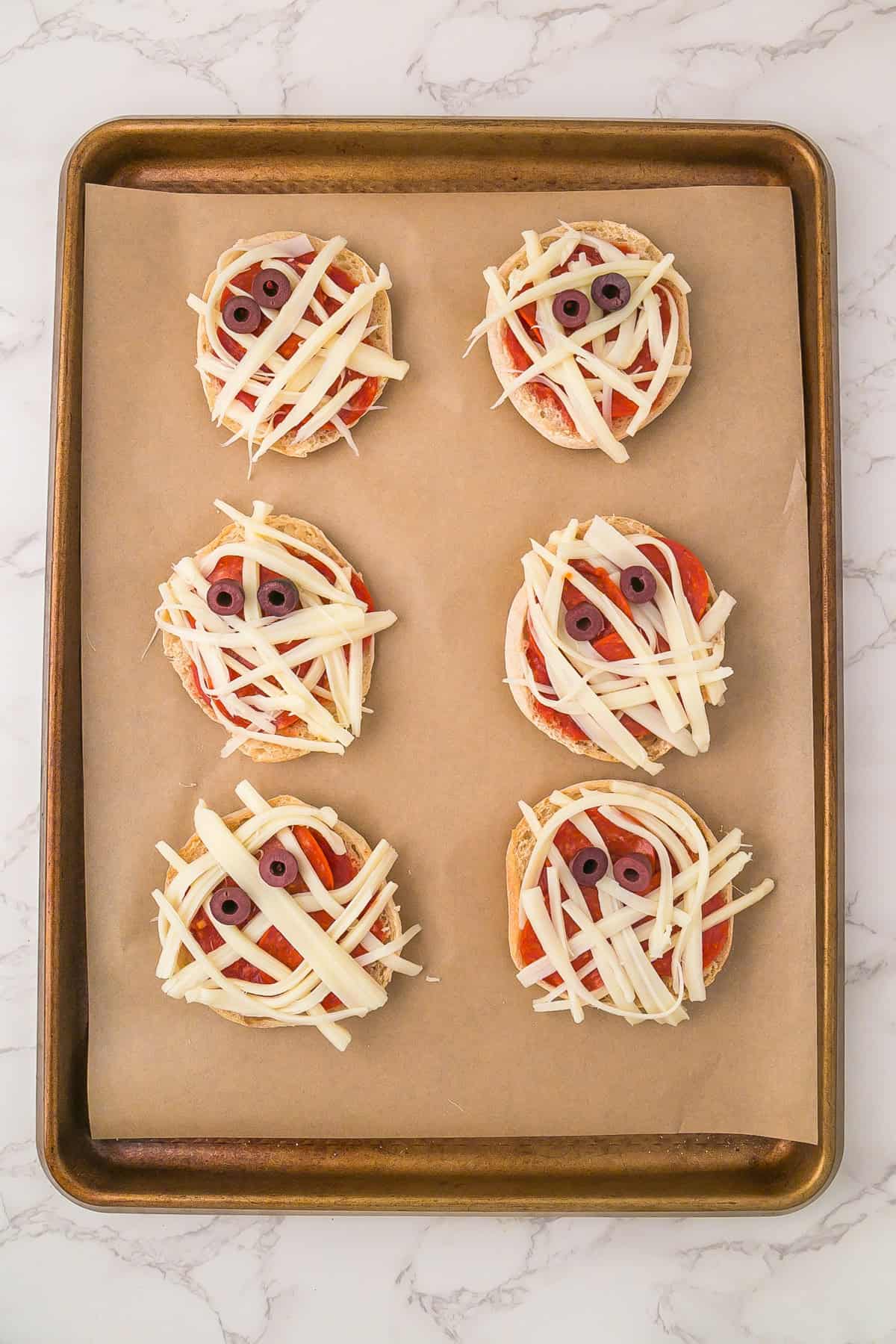 Six unbaked mini mummy pizzas made from English muffins are decorated with strips of cheese and olive slice eyes, all arranged on a baking sheet lined with parchment paper.
