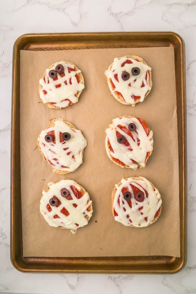 Six cooked mini mummy pizzas made with English muffins, melted cheese, and olive slice eyes are arranged on a parchment-lined baking sheet.
