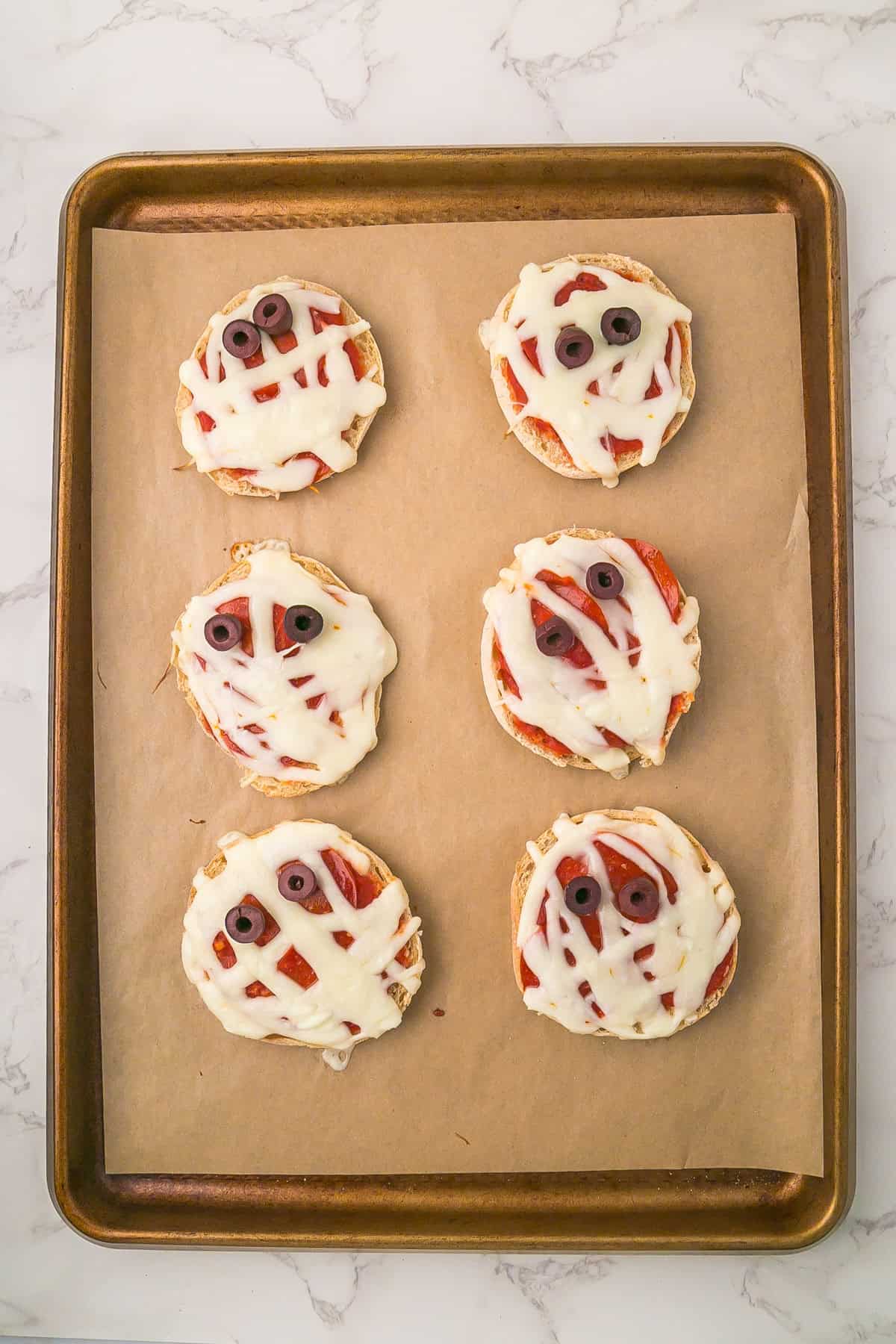 Six cooked mini mummy pizzas made with English muffins, melted cheese, and olive slice eyes are arranged on a parchment-lined baking sheet.