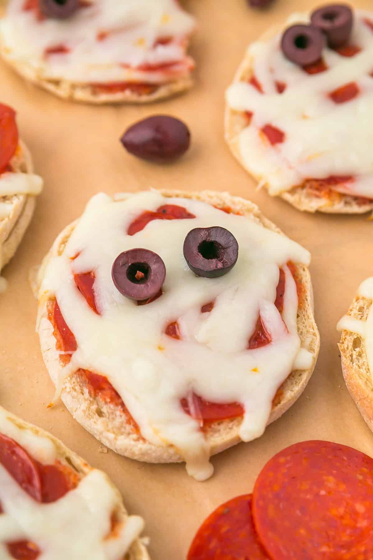 Mini mummy pizzas made with English muffins, topped with melted cheese, pepperoni, and two black olive slices for eyes, arranged on a parchment surface.