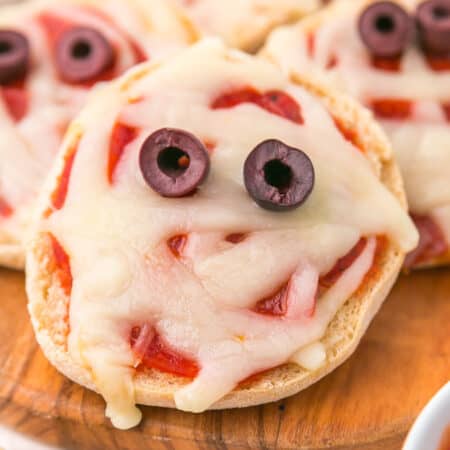 An English muffin pizza topped with melted cheese, two olives for eyes, and pizza sauce, arranged to resemble a mini mu mummy face.