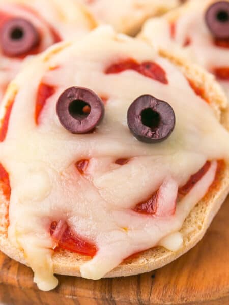 An English muffin pizza topped with melted cheese, two olives for eyes, and pizza sauce, arranged to resemble a mini mu mummy face.