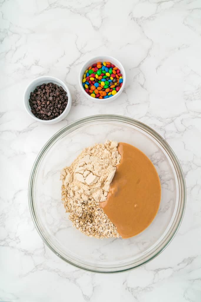A glass bowl with oats, protein powder and peanut butter next to small bowls of mini chocolate chips and M&Ms for monster cookie energy balls.