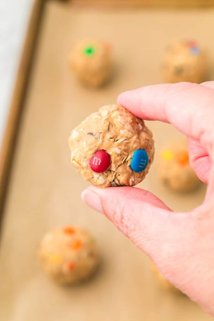 A hand holding a monster cookie energy ball with two M&M candies on it, with more oatmeal balls on a parchment-lined baking sheet in the background.