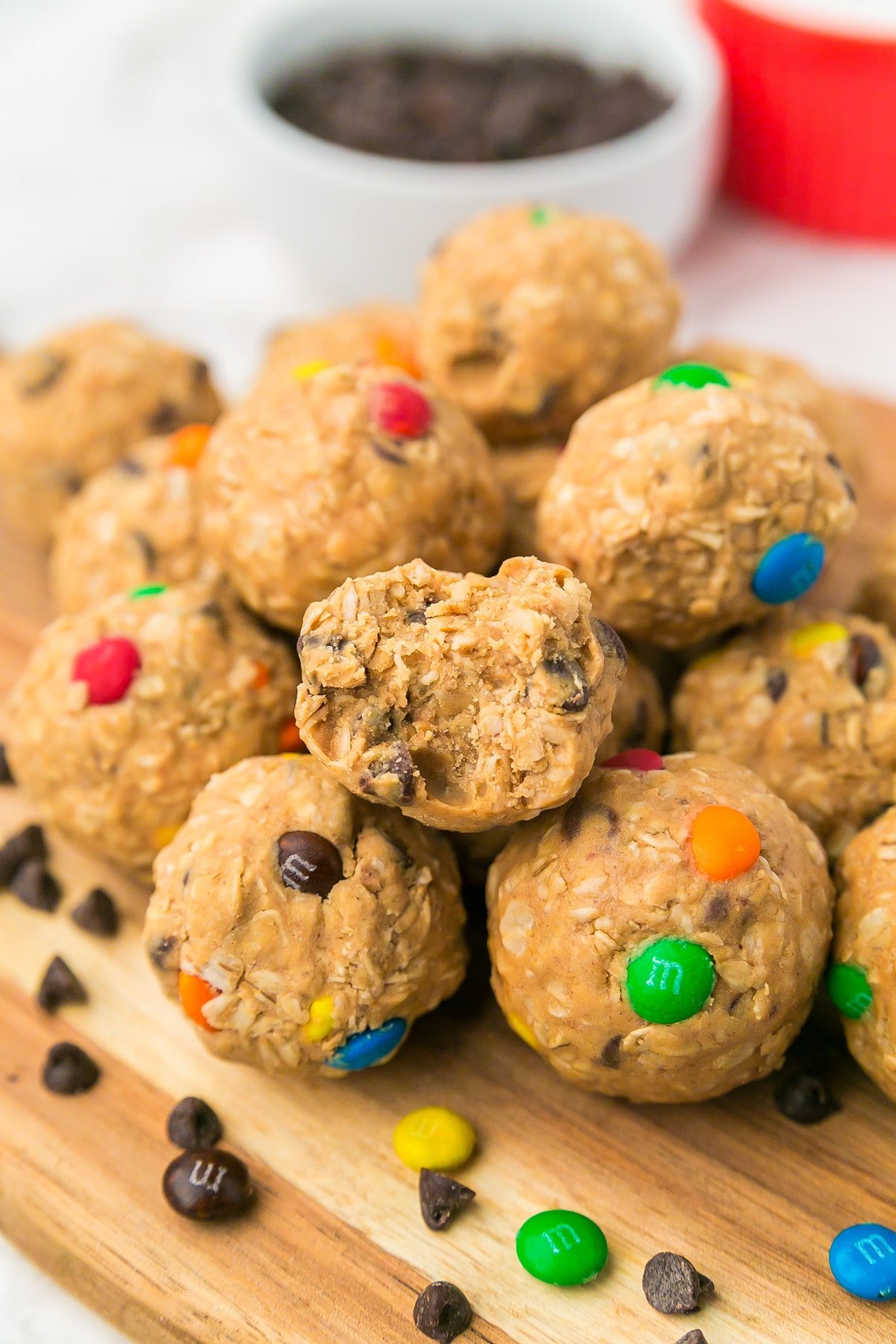 A pile of no-bake monster cookie snack balls with oats, chocolate chips, and colorful candy-coated chocolates on a wooden board with one ball missing a bite.