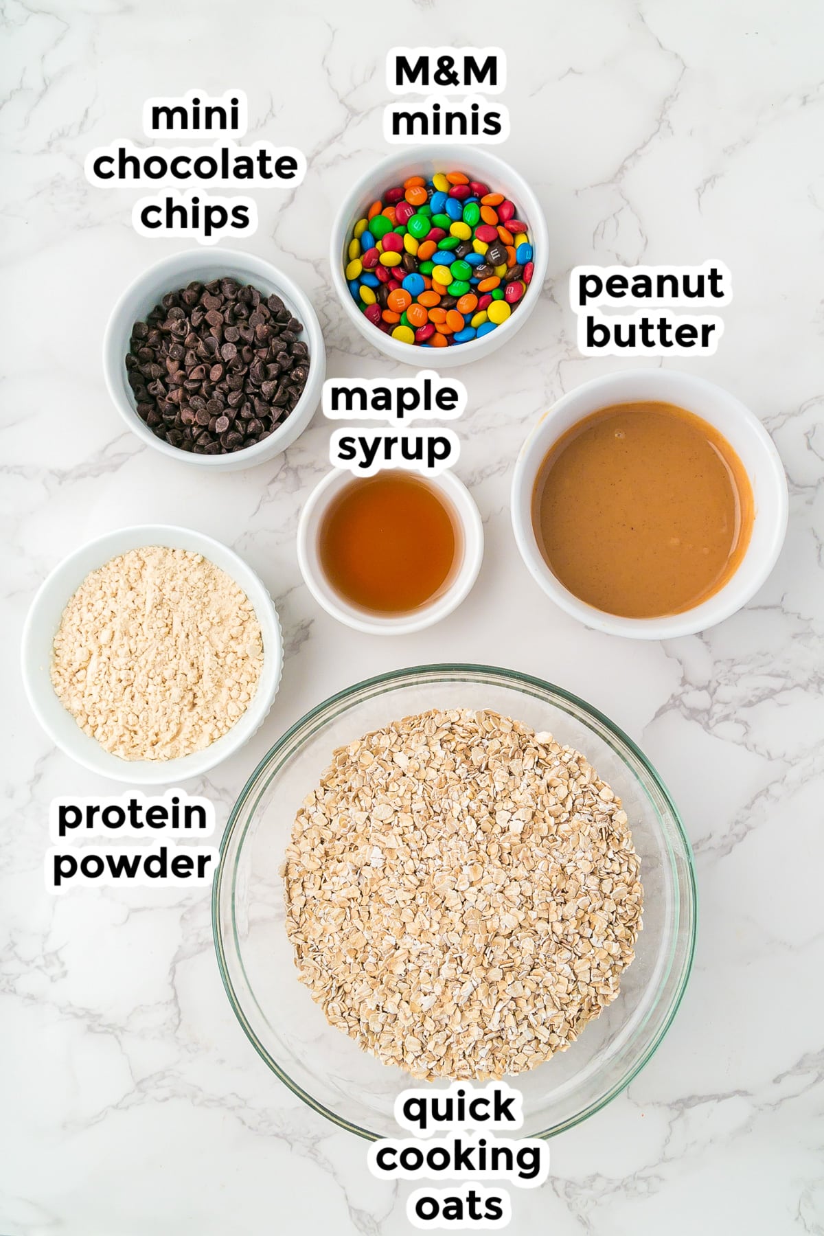 A marble surface displays bowls of quick cooking oats, protein powder, mini chocolate chips, M&M minis, maple syrup, and peanut butter, each labeled with text.