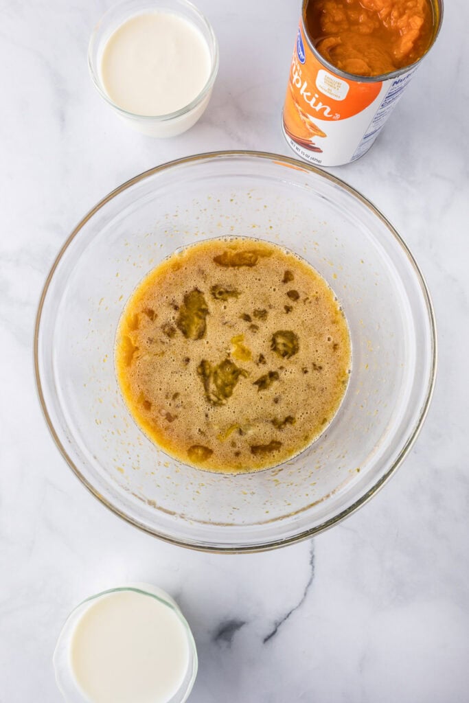A glass bowl containing a mixed egg custard liquid for pumpkin french toast casserole, sits on a marble surface, surrounded by glasses of milk, heavy cream and an open can of pumpkin puree.
