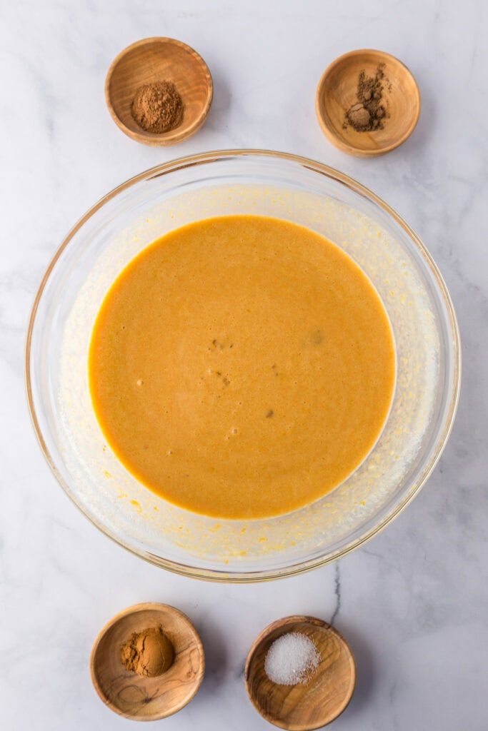 A glass bowl filled with smooth orange custard for pumpkin french toast casserole is surrounded by small wooden bowls of spices and salt.