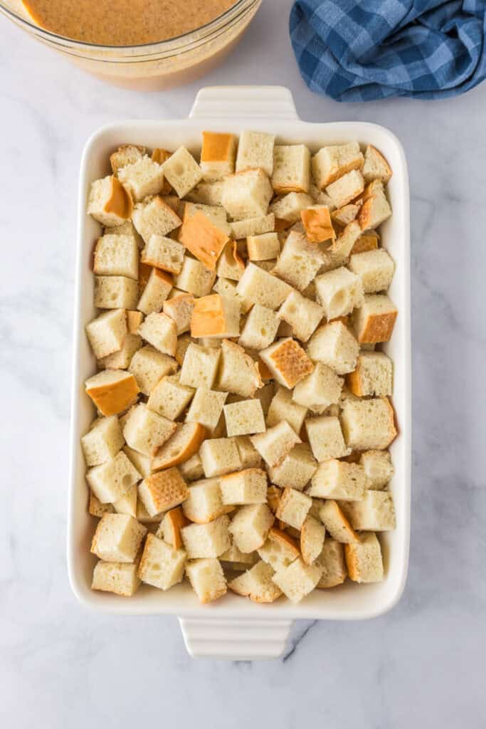 A white baking dish filled with cubed bread for pumpkin French toast casserole sits on a marble surface, next to a blue cloth and a bowl of pumpkin custard mixture.