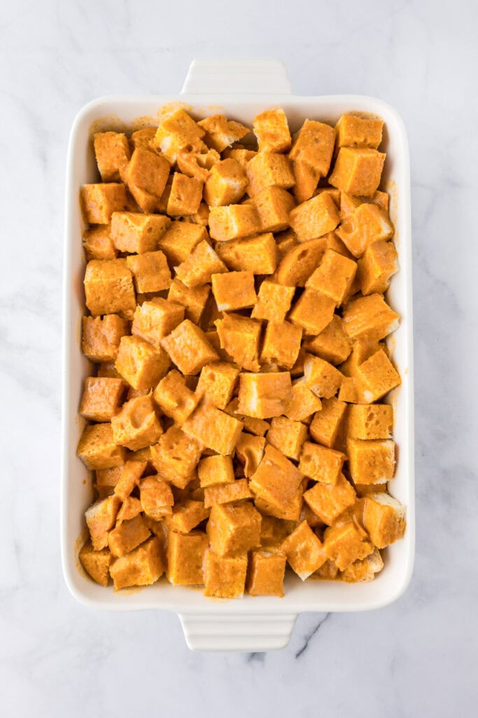 A white baking dish filled with evenly cut cubes of orange pumpkin custard soaked bread coated for pumpkin french toast casserole.