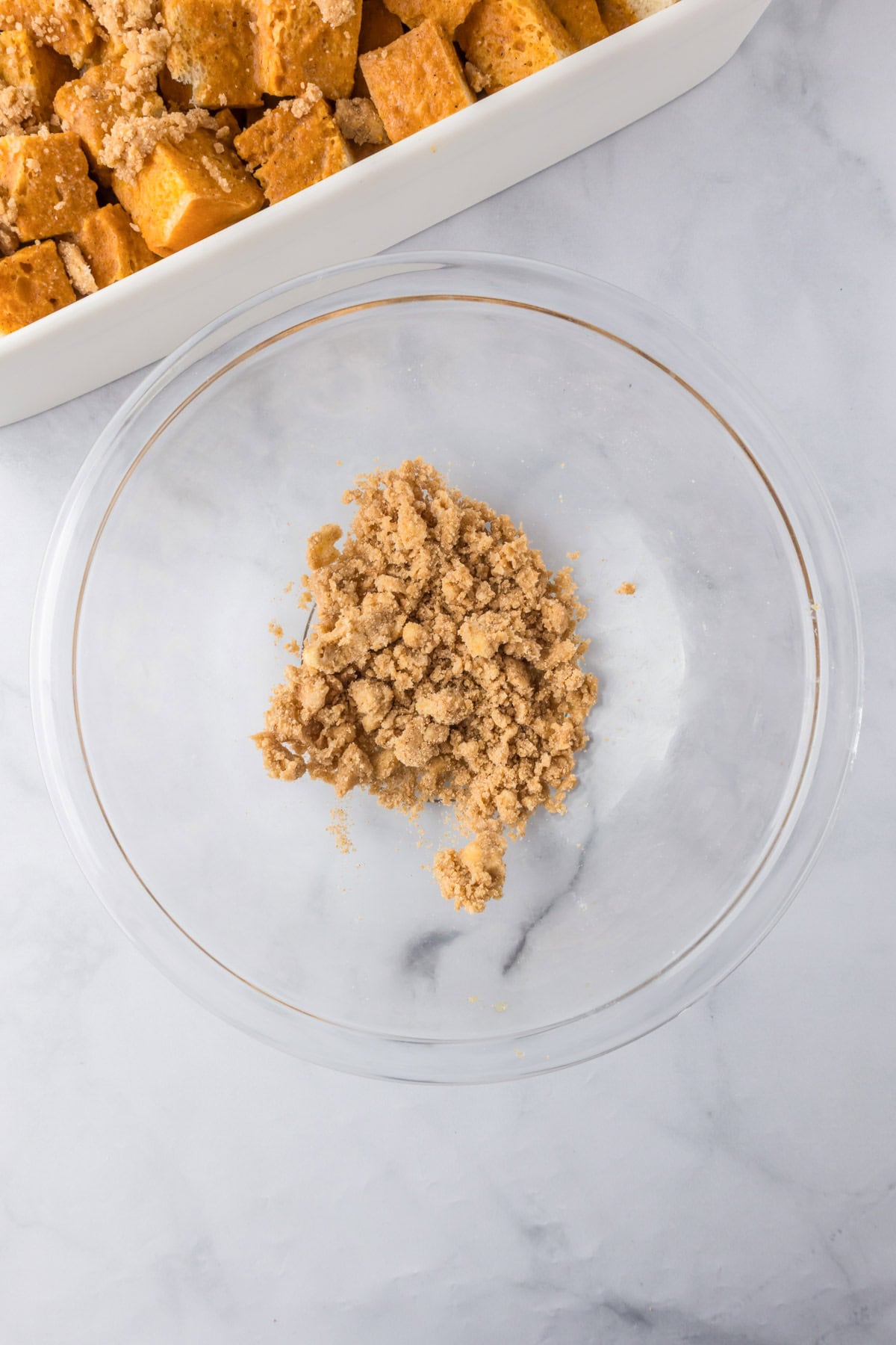 A clear glass bowl containing a small amount of the crumble topping next to a casserole dish full of pumpkin french toast casserole.