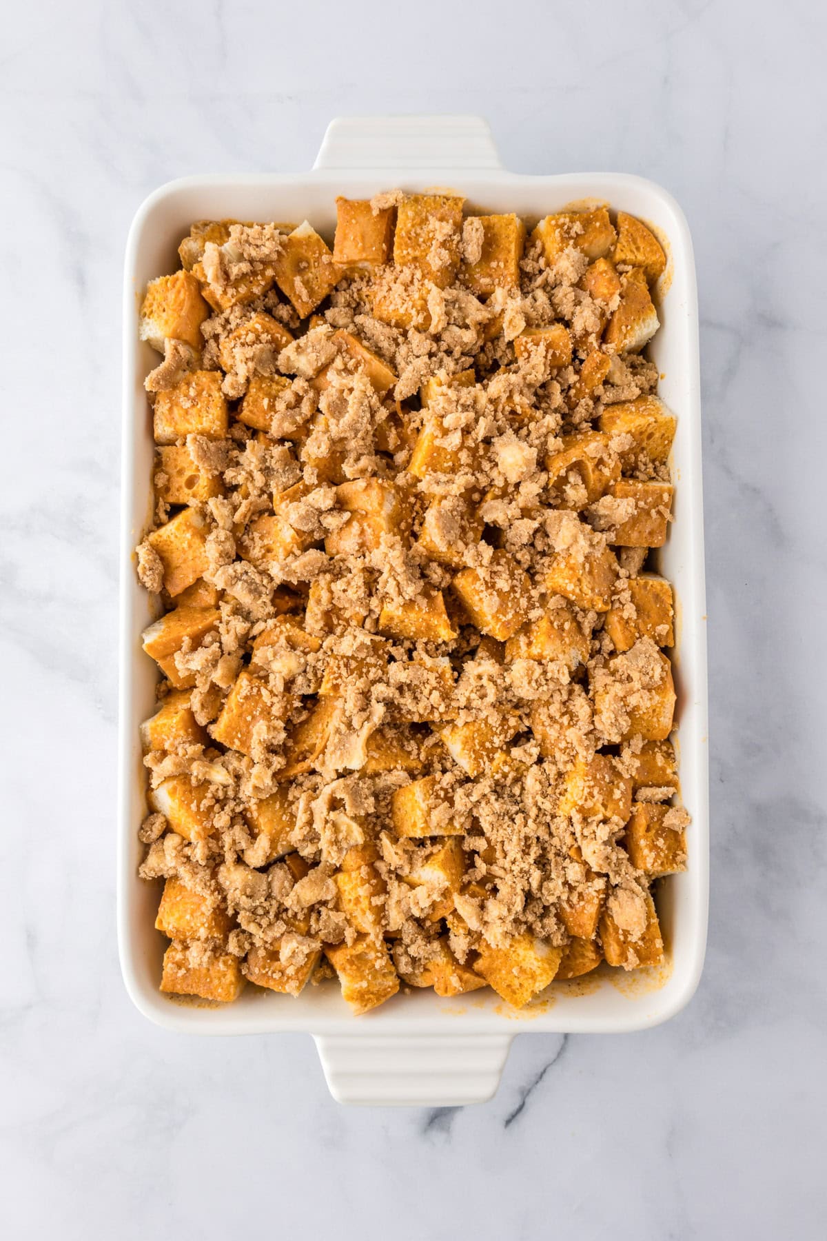 A white rectangular baking dish filled with cubed bread topped with a crumbly brown streusel for a delicious pumpkin french toast casserole before baking.