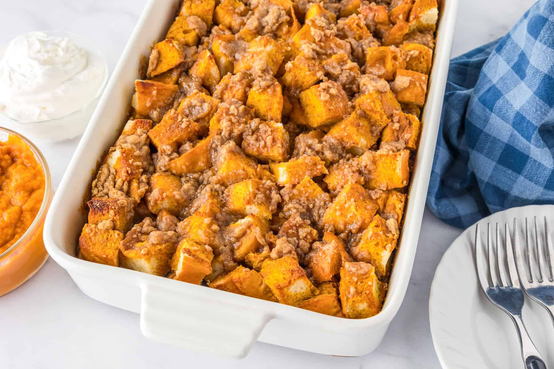 A wide view of a white baking dish filled with pumpkin French toast casserole sits on a table next to a bowl of whipped cream and plates with forks.