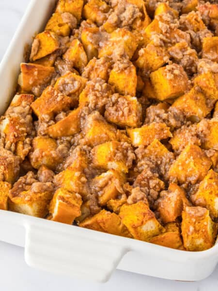 A white baking dish filled with pumpkin French toast casserole topped with a crumbly streusel, served alongside a bowl of whipped cream.