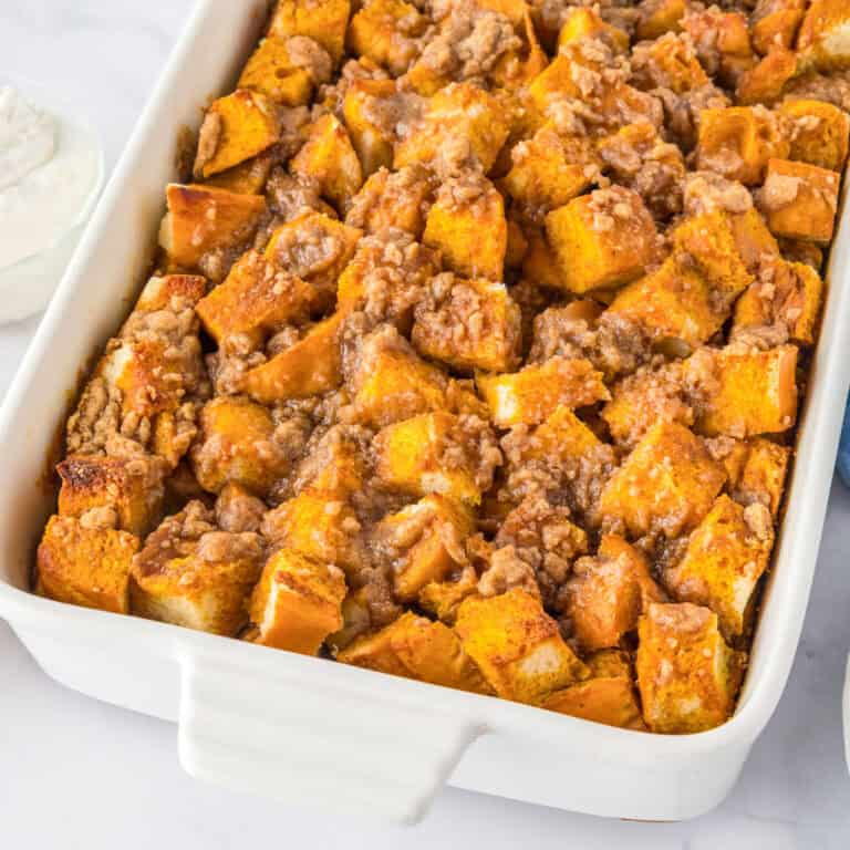 A white baking dish filled with pumpkin French toast casserole topped with a crumbly streusel, served alongside a bowl of whipped cream.