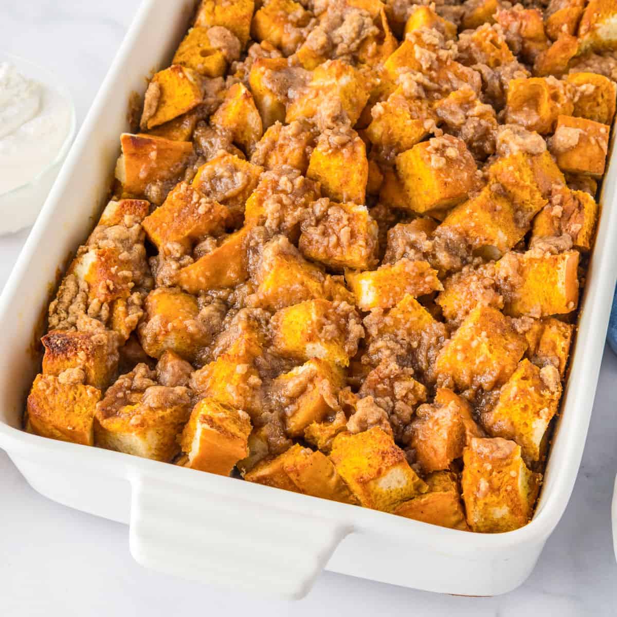 A white baking dish filled with pumpkin French toast casserole topped with a crumbly streusel, served alongside a bowl of whipped cream.