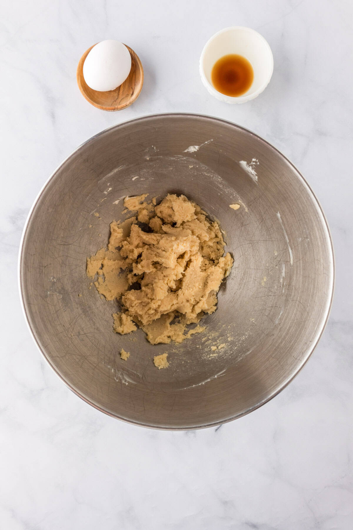 A metal mixing bowl with creamed butter and sugar, ready for making pumpkin oatmeal cookies, sits beside a whole egg in a small dish and a bowl of vanilla extract on a marble countertop.