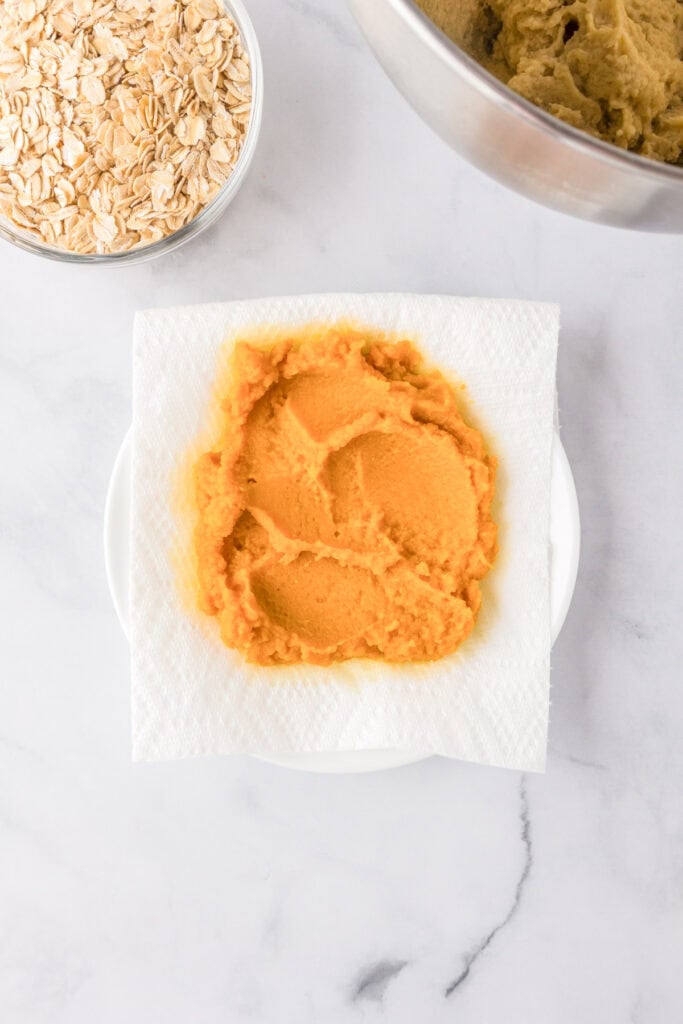 A portion of pumpkin puree on a paper towel atop a white plate, with a bowl of rolled oats and a mixing bowl nearby. Blot the pumpkin so the cookies bake chewy, not cakey.