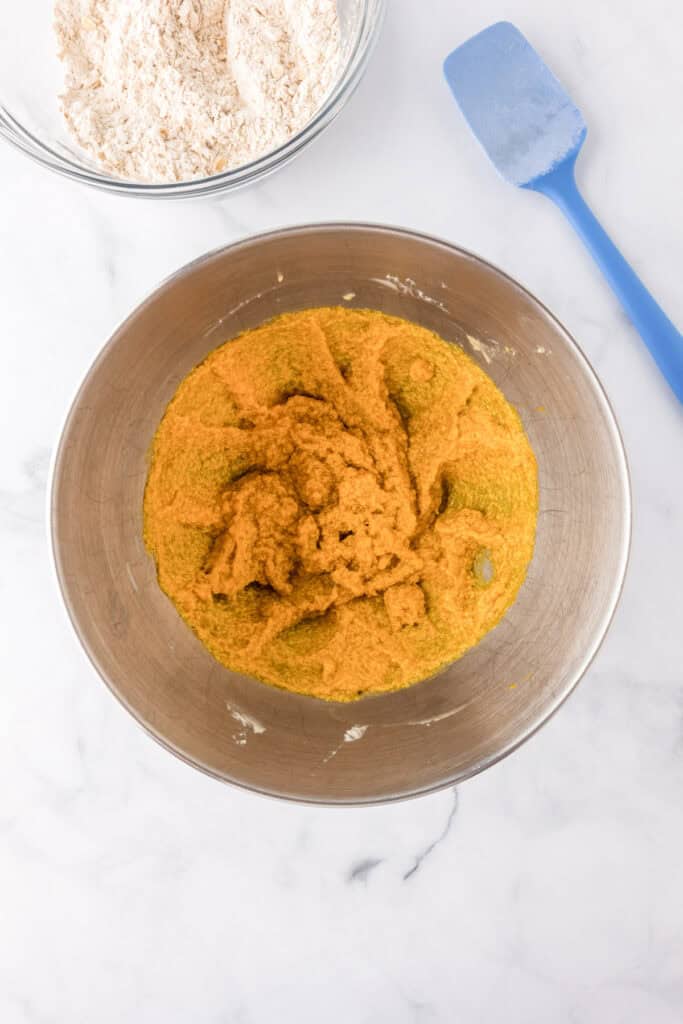 A metal mixing bowl containing a yellowish batter for pumpkin oatmeal cookies sits on a white surface next to a bowl of flour mixture and a blue spatula.