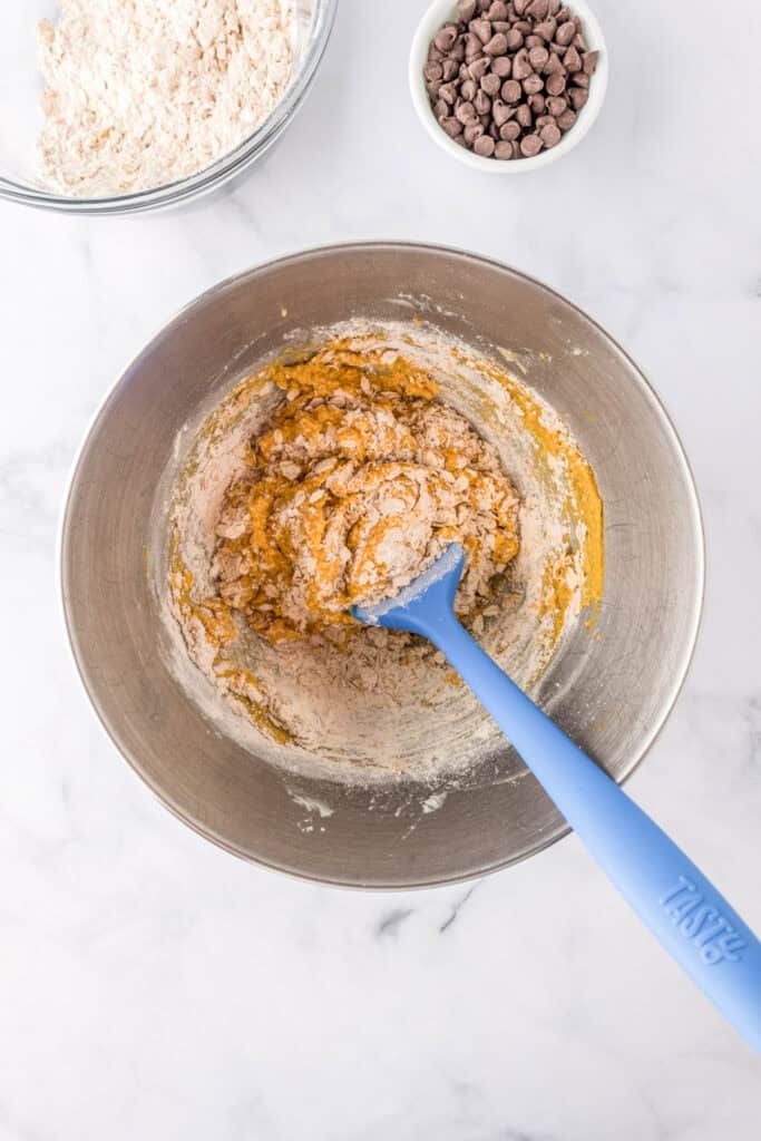 A mixing bowl with pumpkin oatmeal cookie dough being stirred by a blue spatula, surrounded by a bowl of flour and a bowl of chocolate chips on a white surface.