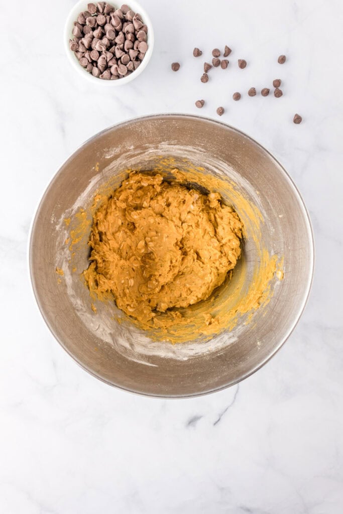 A metal mixing bowl with pumpkin oatmeal cookie dough, accompanied by a small bowl of chocolate chips and some scattered chips on a marble countertop.
