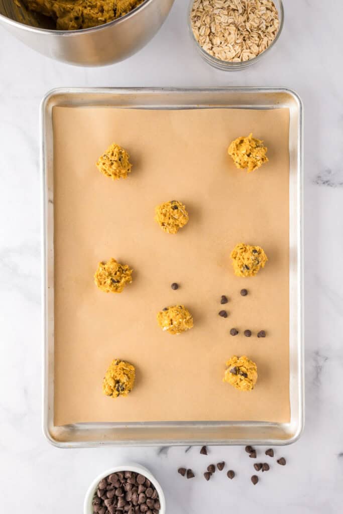 A baking sheet lined with parchment paper holds nine scoops of pumpkin oatmeal cookie dough, each about 2 inches apart. Bowls of oats and chocolate chips sit nearby, with a few chocolate chips scattered on the surface.
