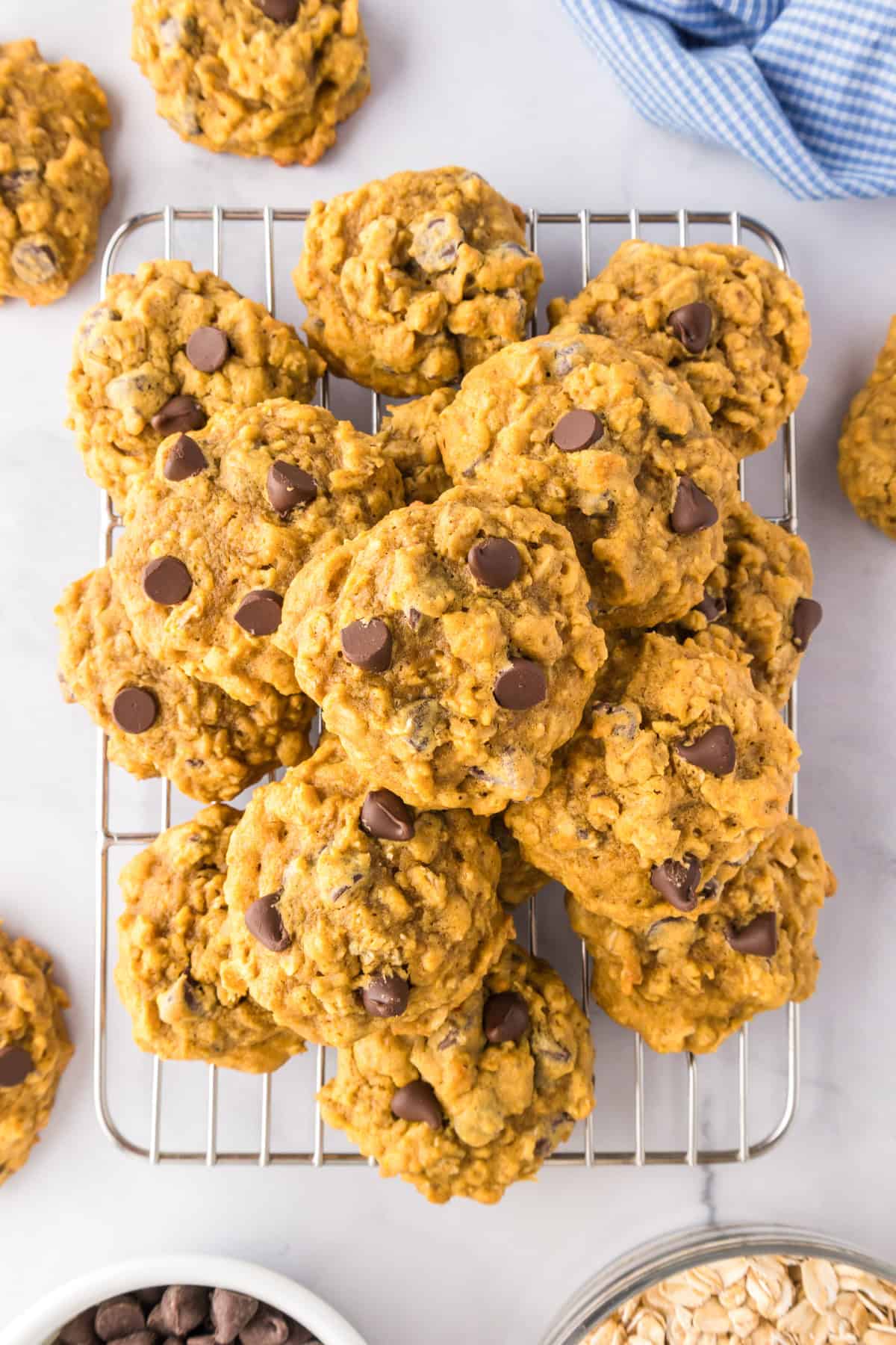 A pile of pumpkin oatmeal cookies stacked on a cooling rack, with a few cookies and chocolate chips scattered nearby.
