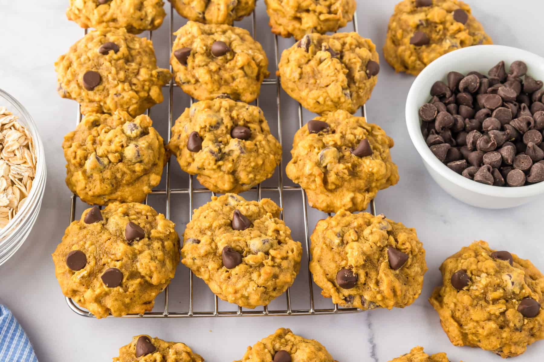 Pumpkin oatmeal cookies packed with chocolate chips cool on a rack, surrounded by a bowl of chocolate chips and oats atop a marble surface.