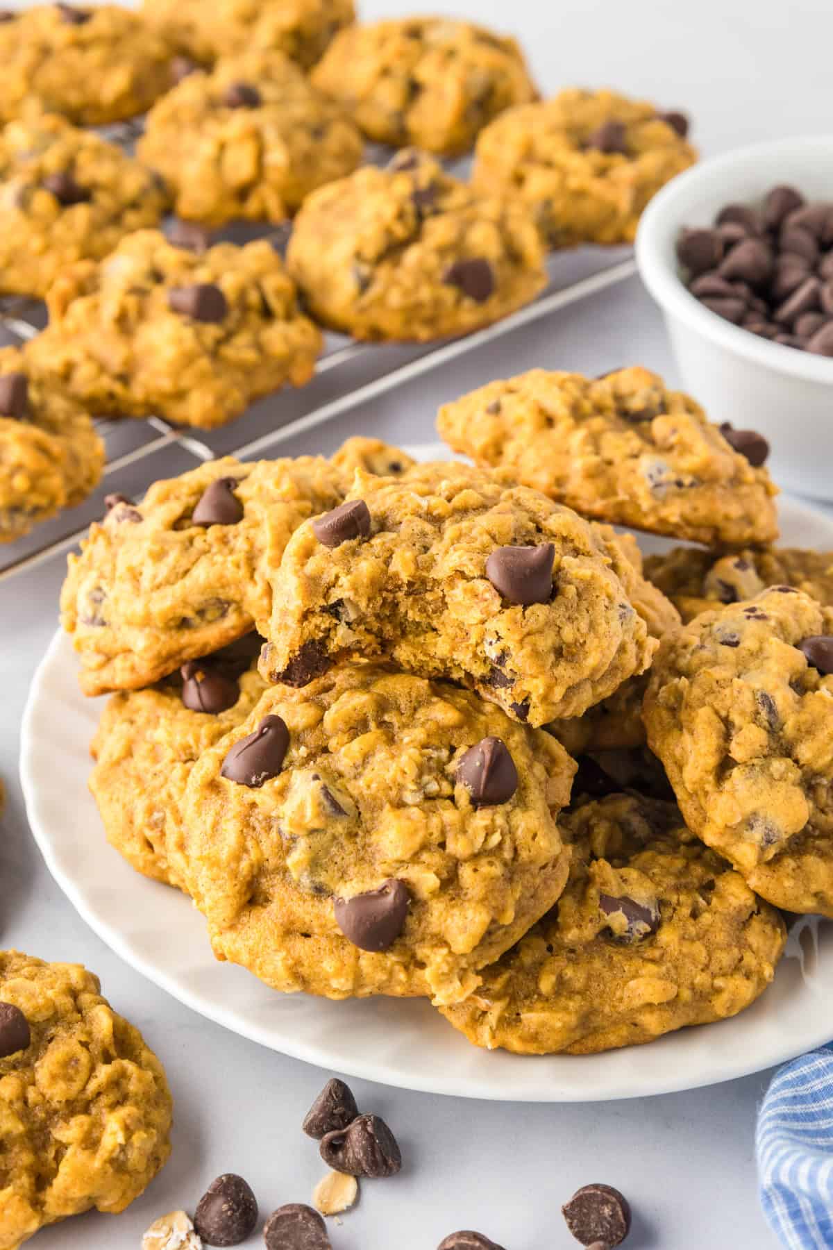 A plate of pumpkin oatmeal chocolate chip cookies is shown, with more pumpkin oatmeal cookies cooling on a wire rack in the background and a bowl of chocolate chips nearby.