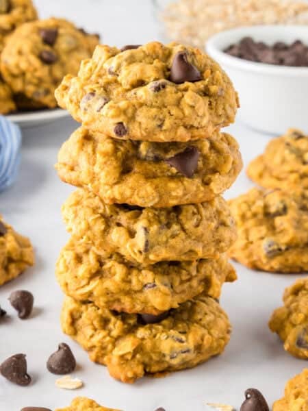 A stack of five pumpkin oatmeal chocolate chip cookies is surrounded by more cookies, chocolate chips, and a bowl of chocolate chips on a white surface.