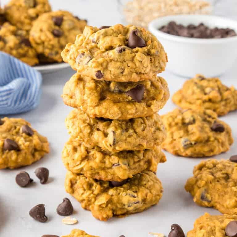A stack of five pumpkin oatmeal chocolate chip cookies is surrounded by more cookies, chocolate chips, and a bowl of chocolate chips on a white surface.
