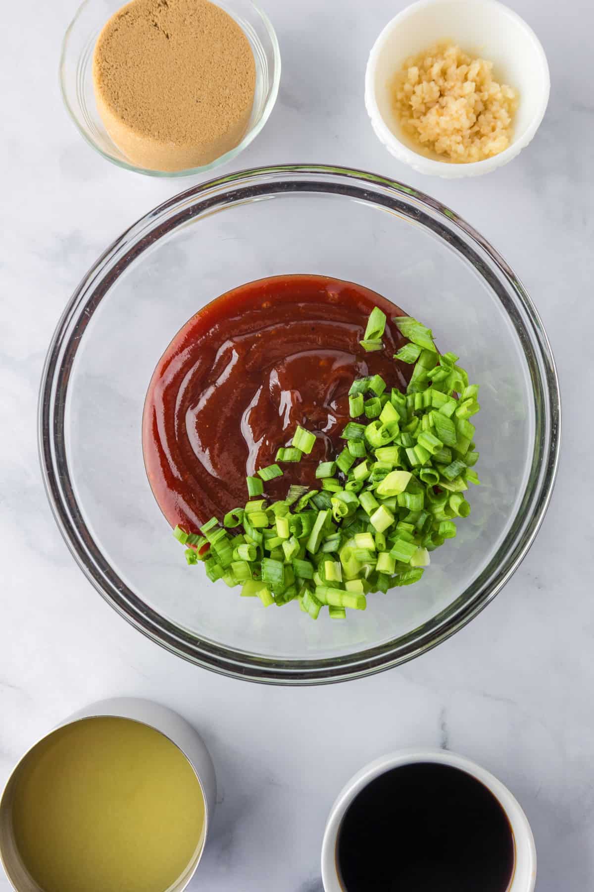 A glass bowl with barbecue sauce and chopped green onions, surrounded by bowls of brown sugar, minced garlic, oil, and soy sauce for Slow Cooker BBQ Hawaiian Roll Chicken Sliders.